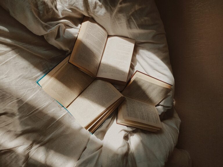 From above opened paper books placed on comfortable bed with white disheveled sheets in daylight